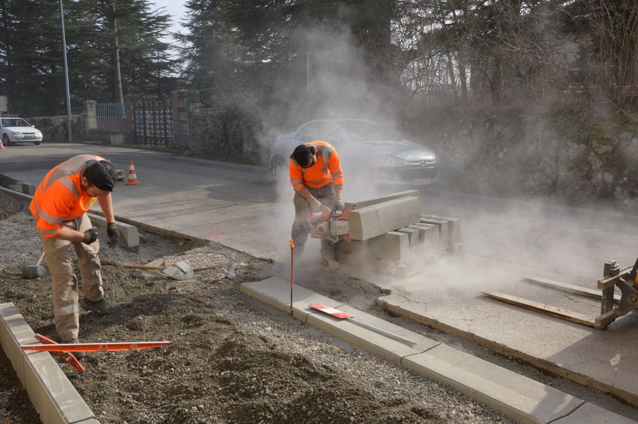 Artisans et métiers du bâtiment à Paris et en Île-de-France Workers cutting concrete on a road with a chainsaw, creating dust and working efficiently. – Carreleur dans les Yvelines (78)